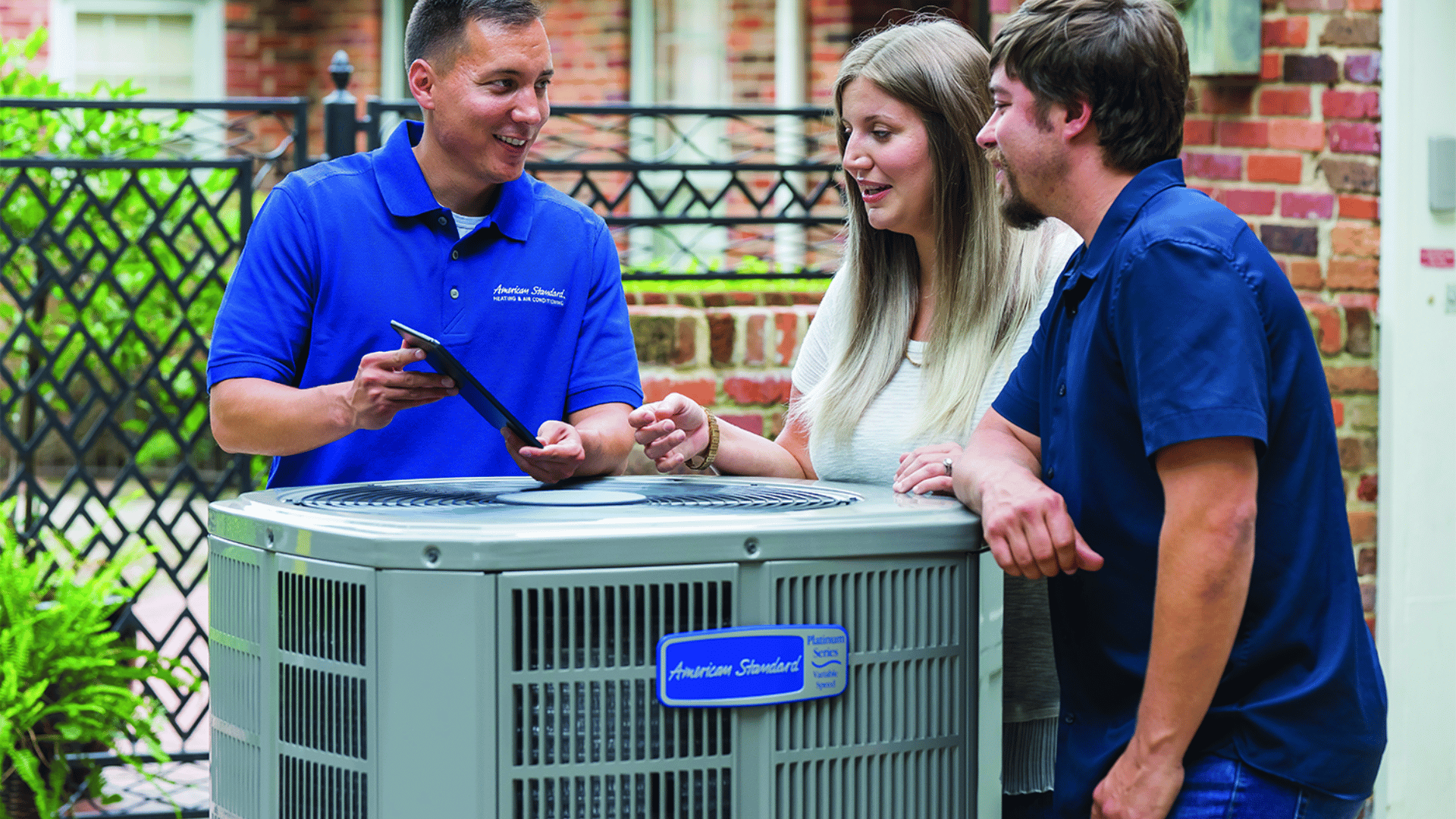 Technician showing information on a tablet to a couple beside an outdoor heat pump unit